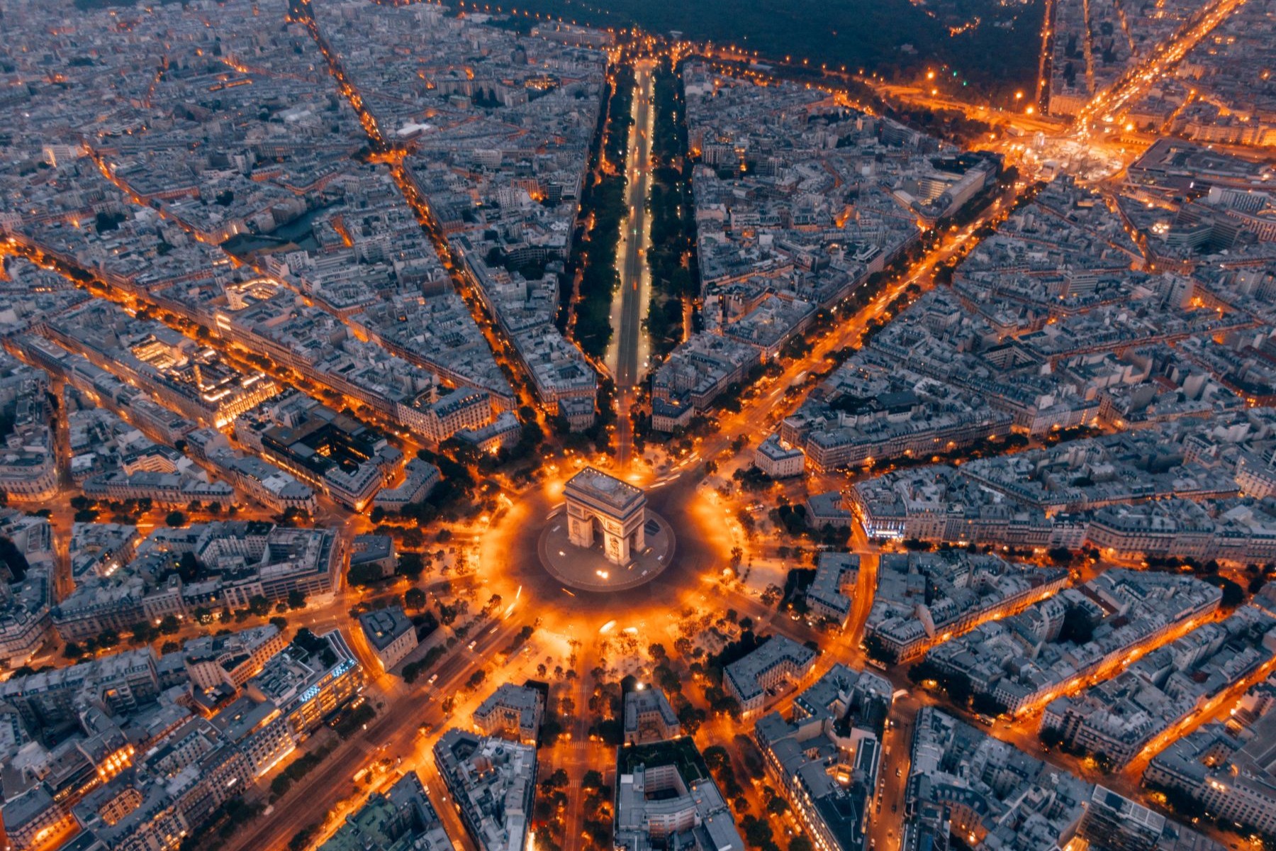 Aerial view of the Arc de Triomphe in Paris at night, surrounded by a circular, illuminated road and radiating city streets.