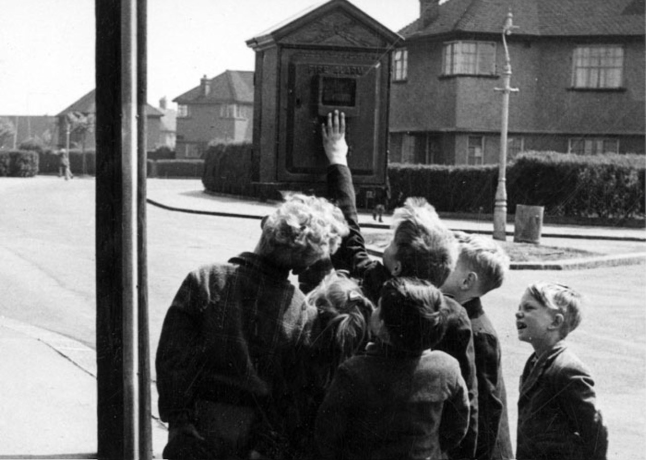 A black and white photograph captures a group of children gathered around a street fire alarm box. One child reaches up to touch the box, while the others look on with curiosity. In the background, houses and trees line the street.