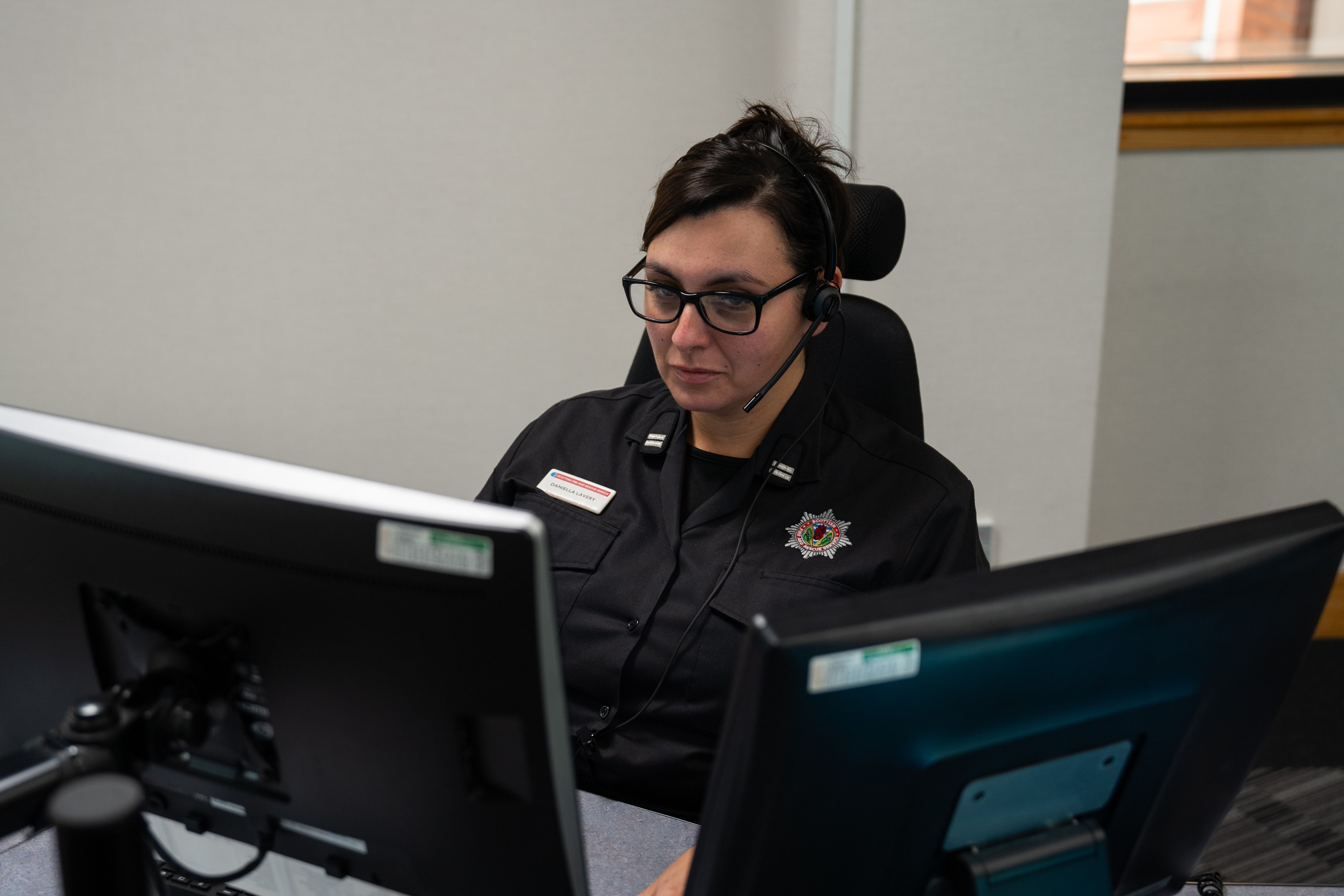 A woman with dark hair wearing glasses and a headset sits at a desk with two computer monitors. She is wearing a black uniform shirt with a badge on the left side of her chest that has a red and white logo. Another emblem is visible on her right shoulder.