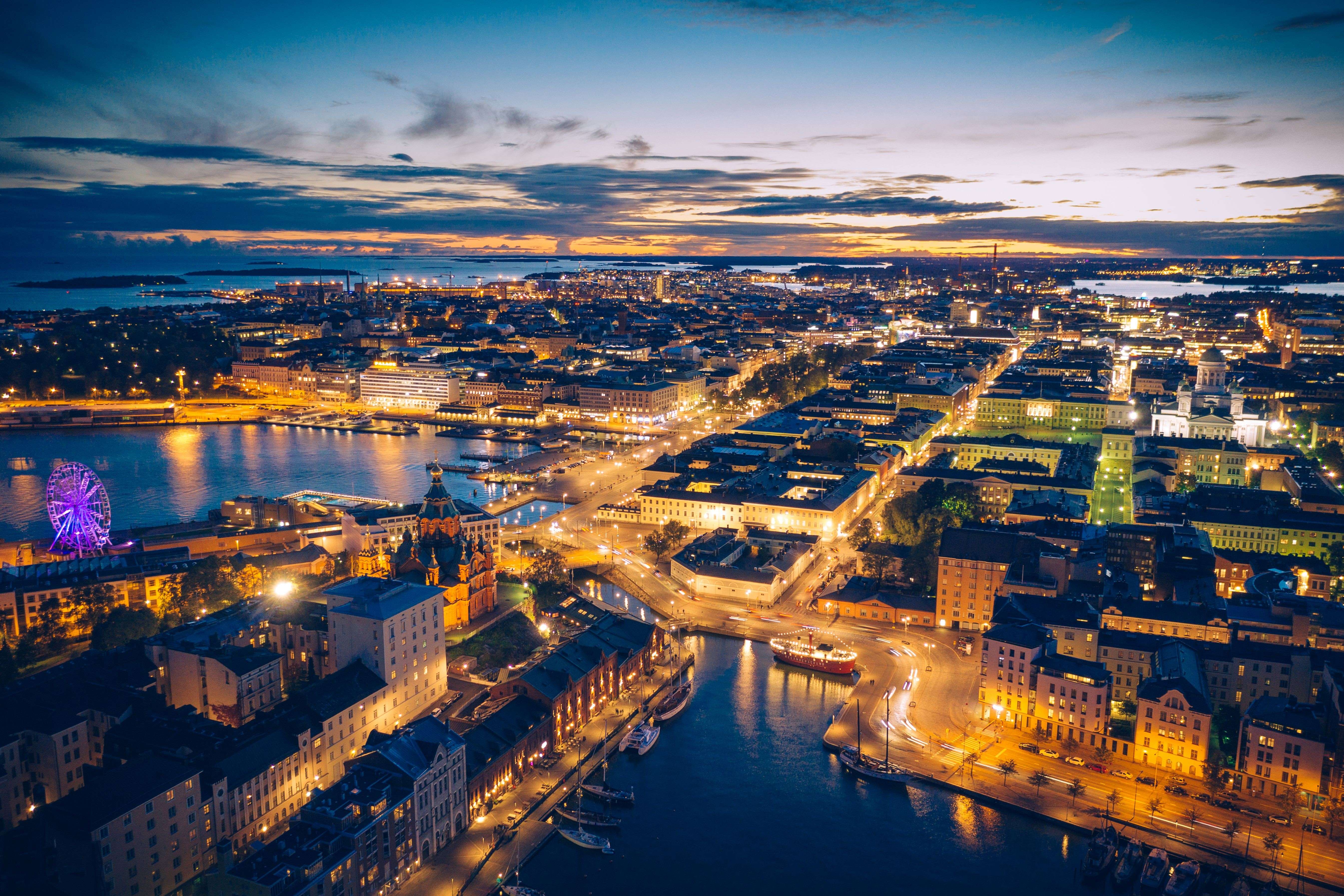 An aerial photograph of a densely built coastal city at twilight. The scene is dominated by a harbor and river running through the city center, with buildings intensely lit in warm orange and yellow. A bright purple-lit Ferris wheel is visible on the left edge, and the horizon shows the last streaks of an orange sunset over the water and distant landmasses. (This city appears to be Helsinki, Finland).