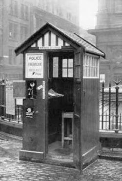 police-box A black and white image displays a police box, resembling a small, wooden hut with a peaked roof and horizontal wooden planks on its side. The front of the box features a window and an open door, revealing a stool inside. On the exterior, a sign reads "POLICE FIRE BRIGADE AMBULANCE PHONE." To the left of the door, a wall-mounted telephone is visible, alongside another sign that is partially obscured but appears to be a newspaper. The police box is situated on a paved surface next to a metal fence, with buildings in the background.
