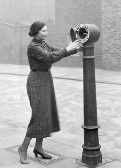 street-fire-alarm A vintage black and white photograph captures a woman in a plaid dress, positioned beside a street fire alarm. The woman has dark hair styled in a bun and wears low heels. Her hands are extended towards the alarm box, which is mounted on a tall, dark pole. The alarm box has the words "FIRE CALL" visible on its side. The setting appears to be a city street, with buildings in the background.