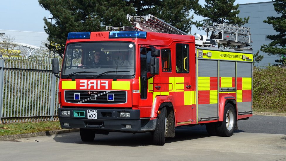 A red and yellow fire engine is driving on a paved road, possibly an industrial or commercial area, with a chain link fence to the left and trees and buildings in the background. The vehicle is a Volvo model and has the license plate AY53 AXD. The side of the vehicle says "FIRE AND RESCUE SERVICE." The front of the vehicle has the word "FIRE" written in reverse.